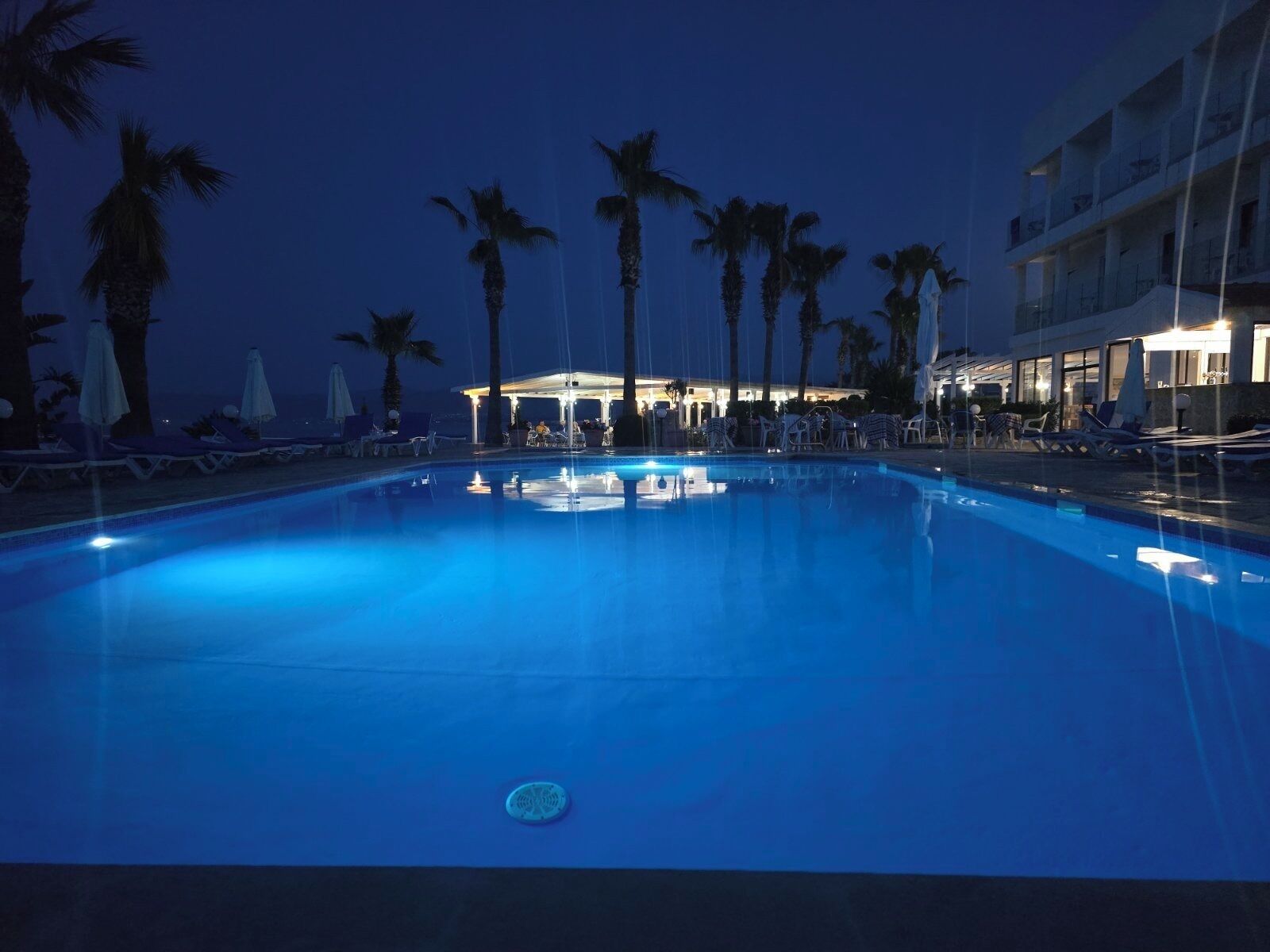 Una piscina con lettini e ombrelloni, circondata da palme e un edificio illuminato.