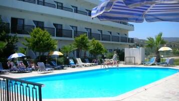 Un área de piscina con sillas de descanso y sombrillas, un edificio con balcones y palmeras.