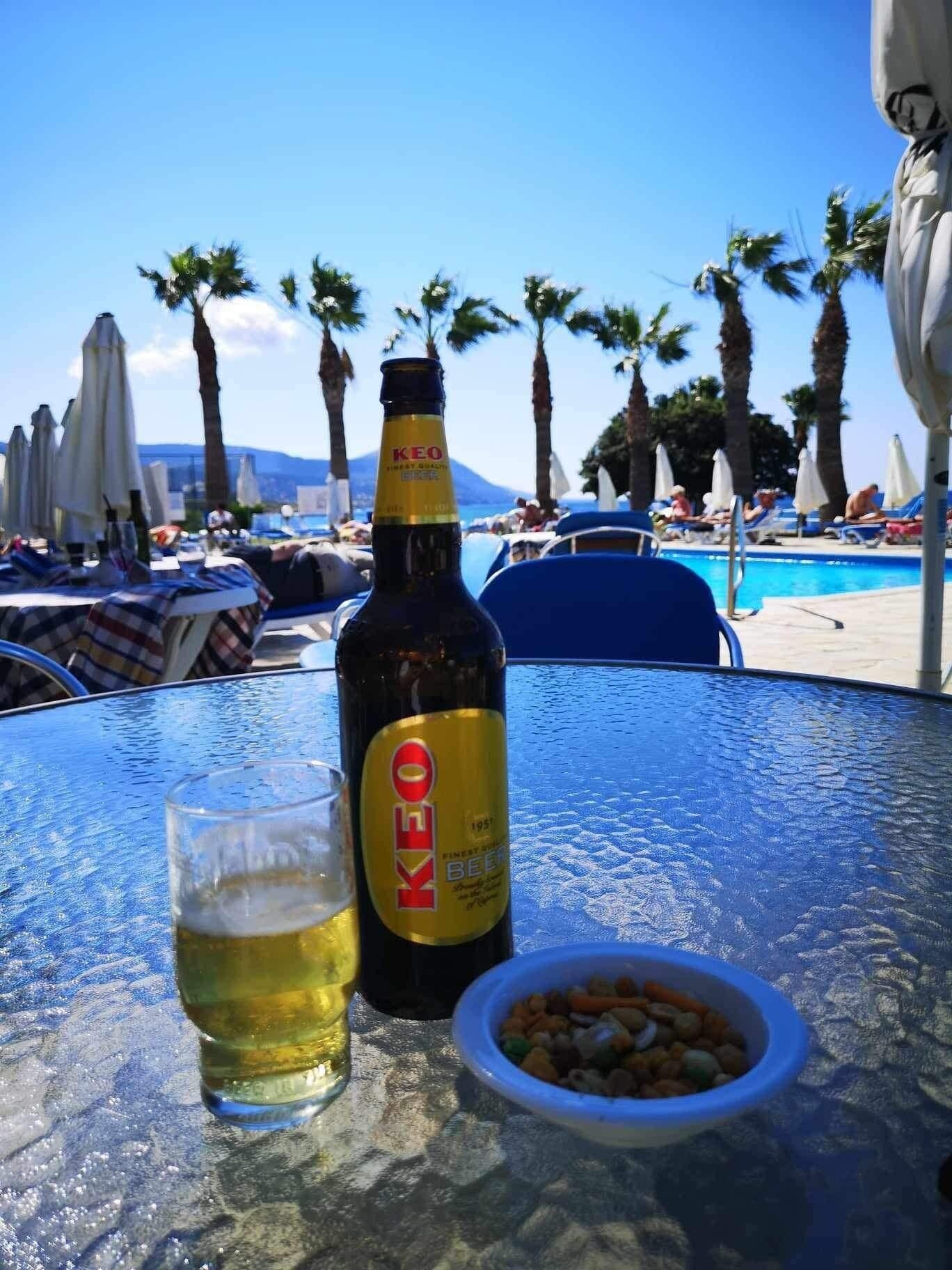 A bottle of KEO beer, a glass of beer, and a bowl of snacks on a table with a pool and palm trees in the background.