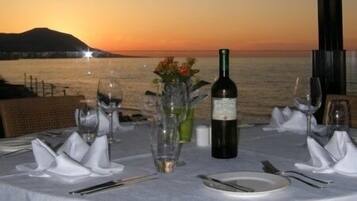 A table set for dinner with a bottle of wine, glasses, and a vase with flowers, overlooking a sunset over the sea.