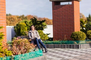 Una mujer sentada en un banco en un patio con un muro de ladrillo y follaje al fondo.