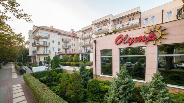 A hotel with a sun logo, multiple balconies, and a well-maintained garden. - Olymp Spa (Kolobrzeg)