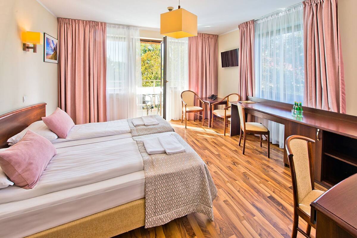 A hotel room with a large bed, a desk, and a chair. There is a window with curtains, a television, and a lamp.