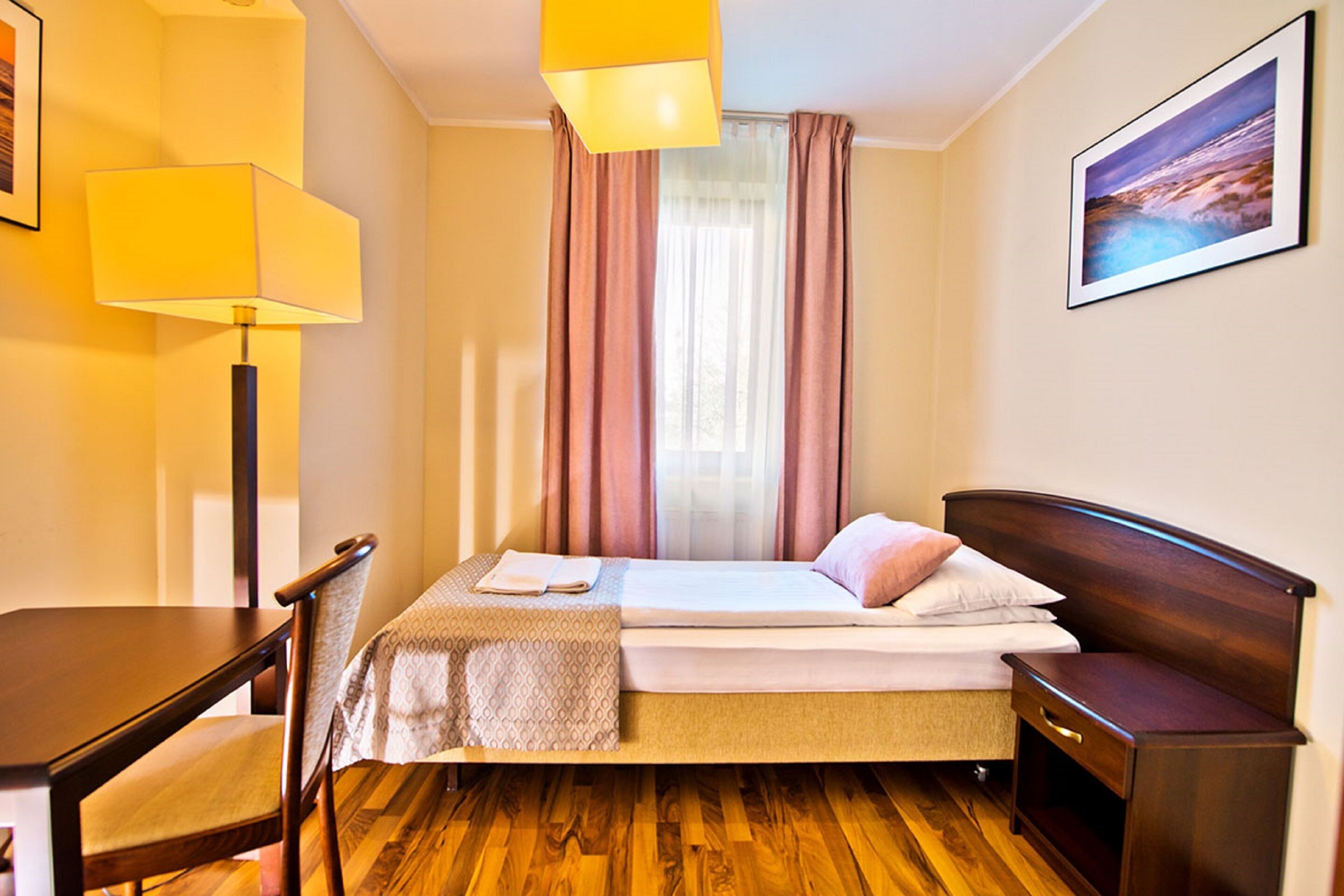 A hotel room with a bed, bedside table, chair, lamp, and a painting on the wall.
