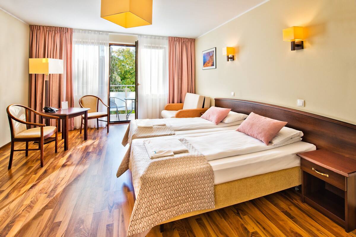 A hotel room with two beds, a desk, chairs, a small table, and a balcony with curtains.