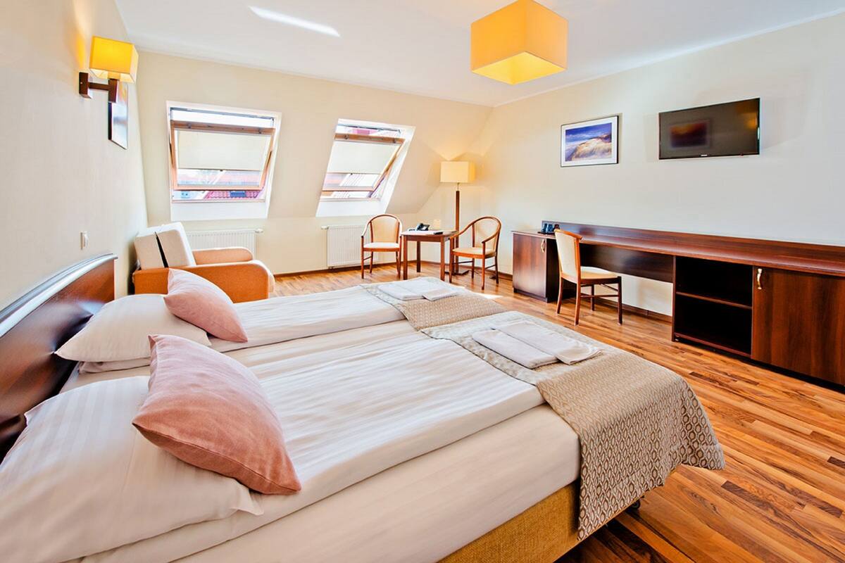 A hotel room with two beds, a desk, and a chair. There are two windows and a TV on the wall.