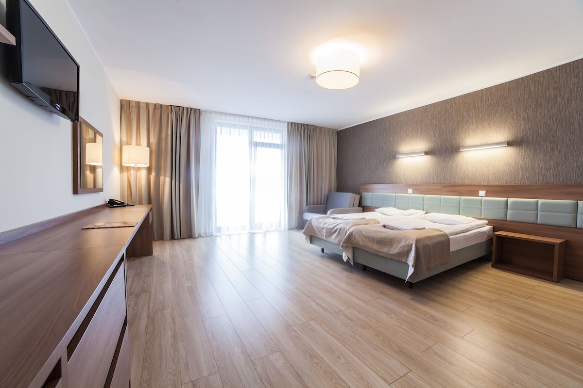 Ein modernes Hotelzimmer mit Holzboden, einem Bett mit Kopfteil, einem Schreibtisch und einem Flachbildfernseher sowie einem großen Fenster mit Vorhängen.