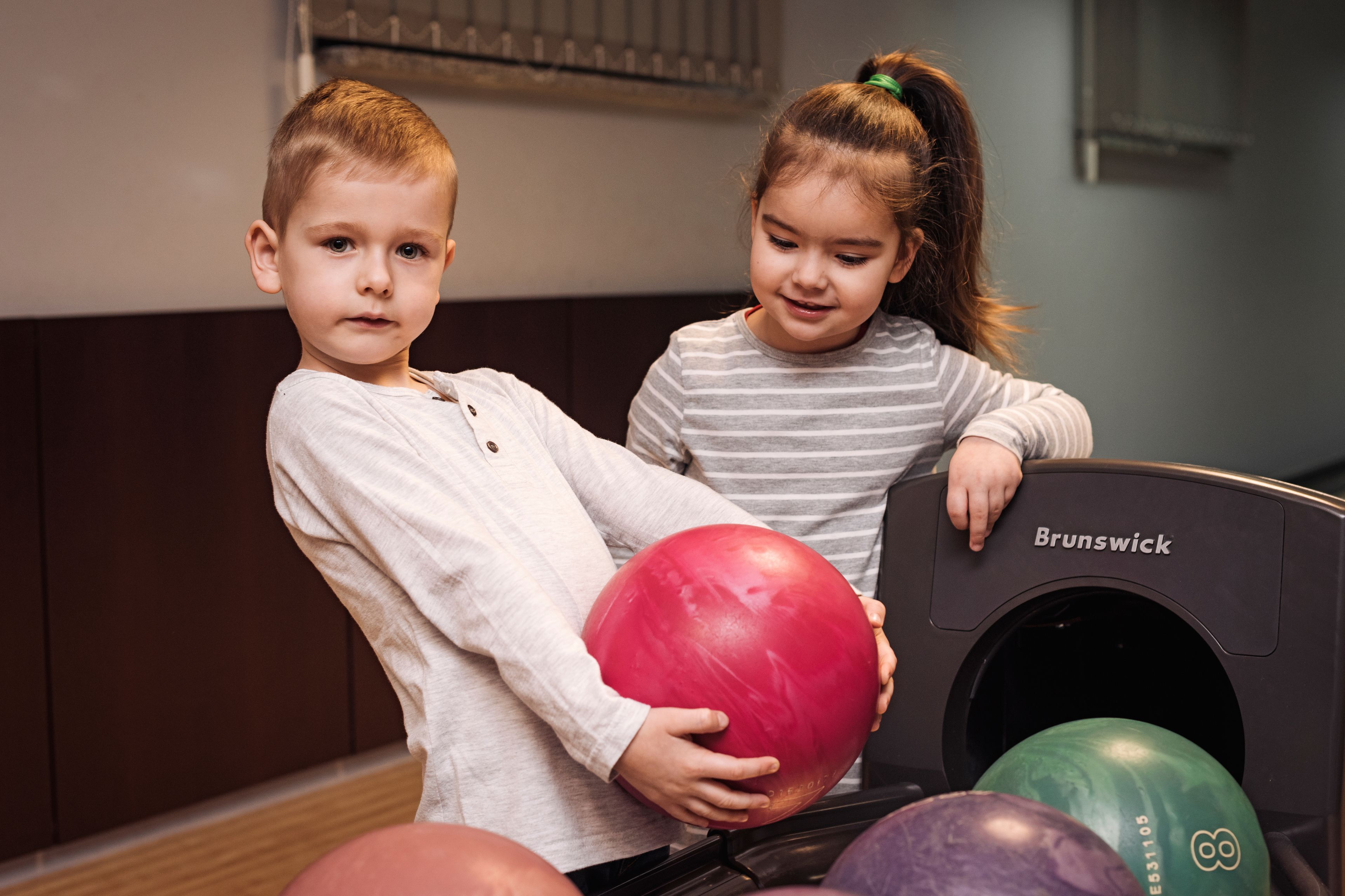 Zwei Kinder tragen Bowlingkugeln in einer Kegelbahn.