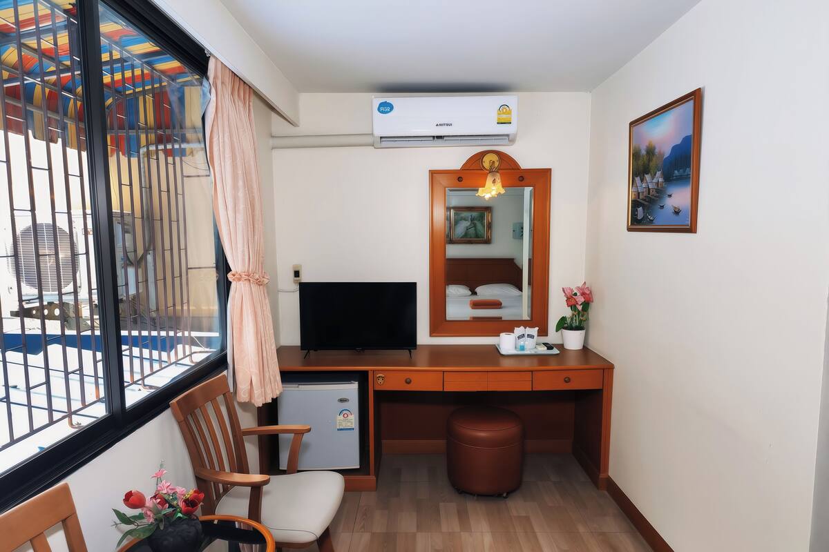 A hotel room with a wooden desk, a television, a small refrigerator, a chair, a potted plant, and a window with a view of a playground.