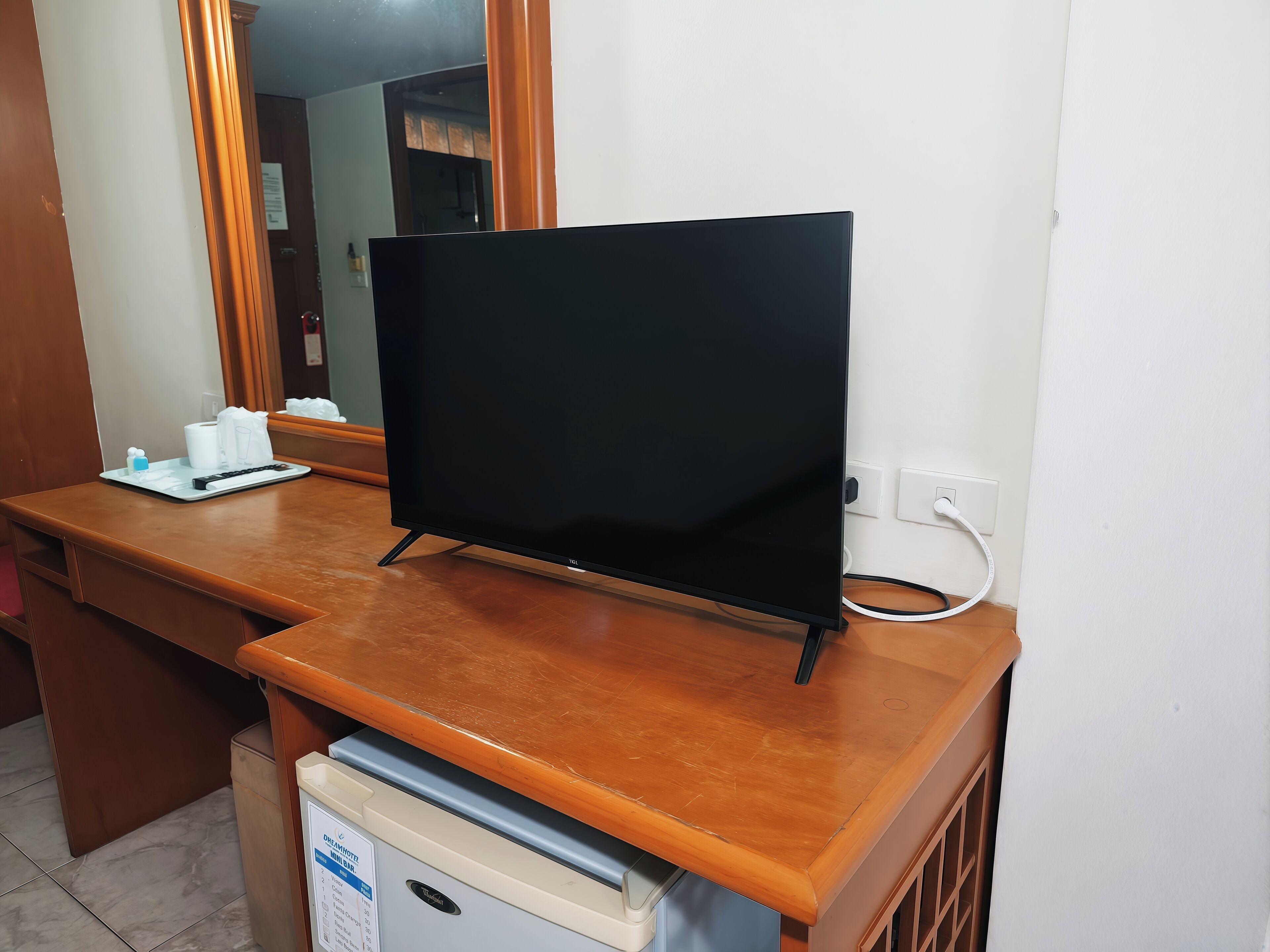 A hotel room with a wooden desk, a flat-screen TV, a mini fridge, and a mirror.