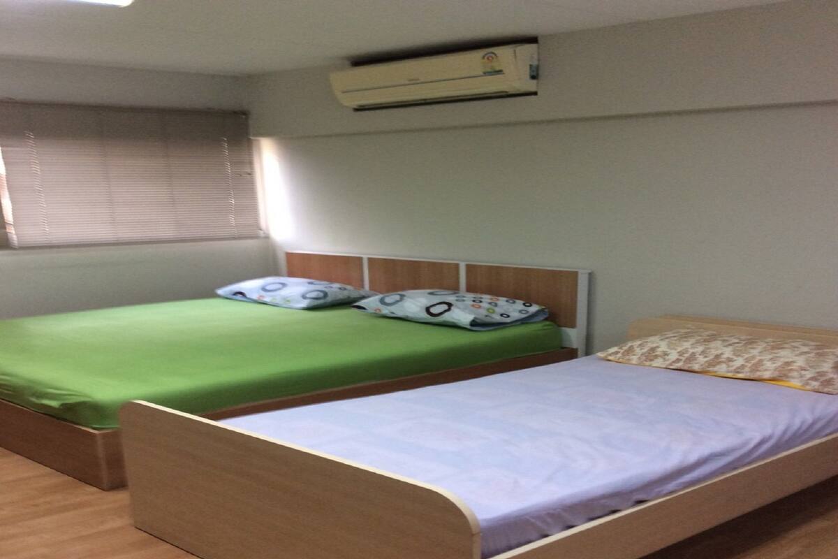 Habitació amb dos llits individuals, un amb estores verdes i l'altre amb estores morades, amb almoinades estampades. Hi ha una finestra amb persianes, un aparell d'aire condicionat sobre els llits i un terra de fusta.