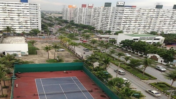 Una cancha de tenis rodeada de palmeras y un estacionamiento, con edificios altos al fondo.