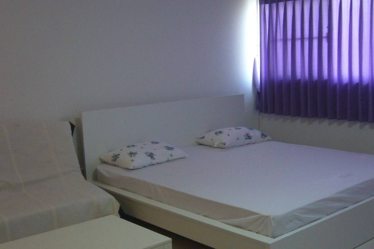 Habitació senzilla i ordenada amb un llit individual, dos coixins, una tauleta de nit i una finestra amb cortines morades.