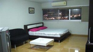 Pequeña habitación amoblada con una cama, una silla negra, una mesa blanca y una ventana con vista a la ciudad de noche.