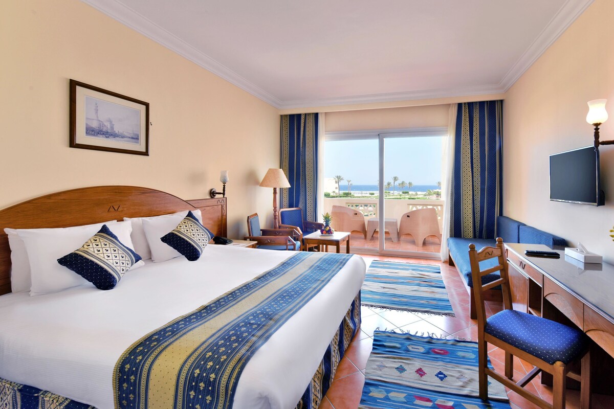 A hotel room with a large bed, a desk, a chair, a TV, and a balcony with a view of the ocean.