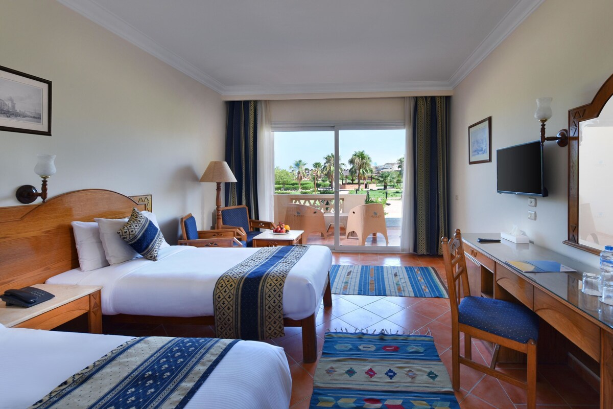 A hotel room with two beds, a desk, a chair, a TV, and a balcony with a view of palm trees.