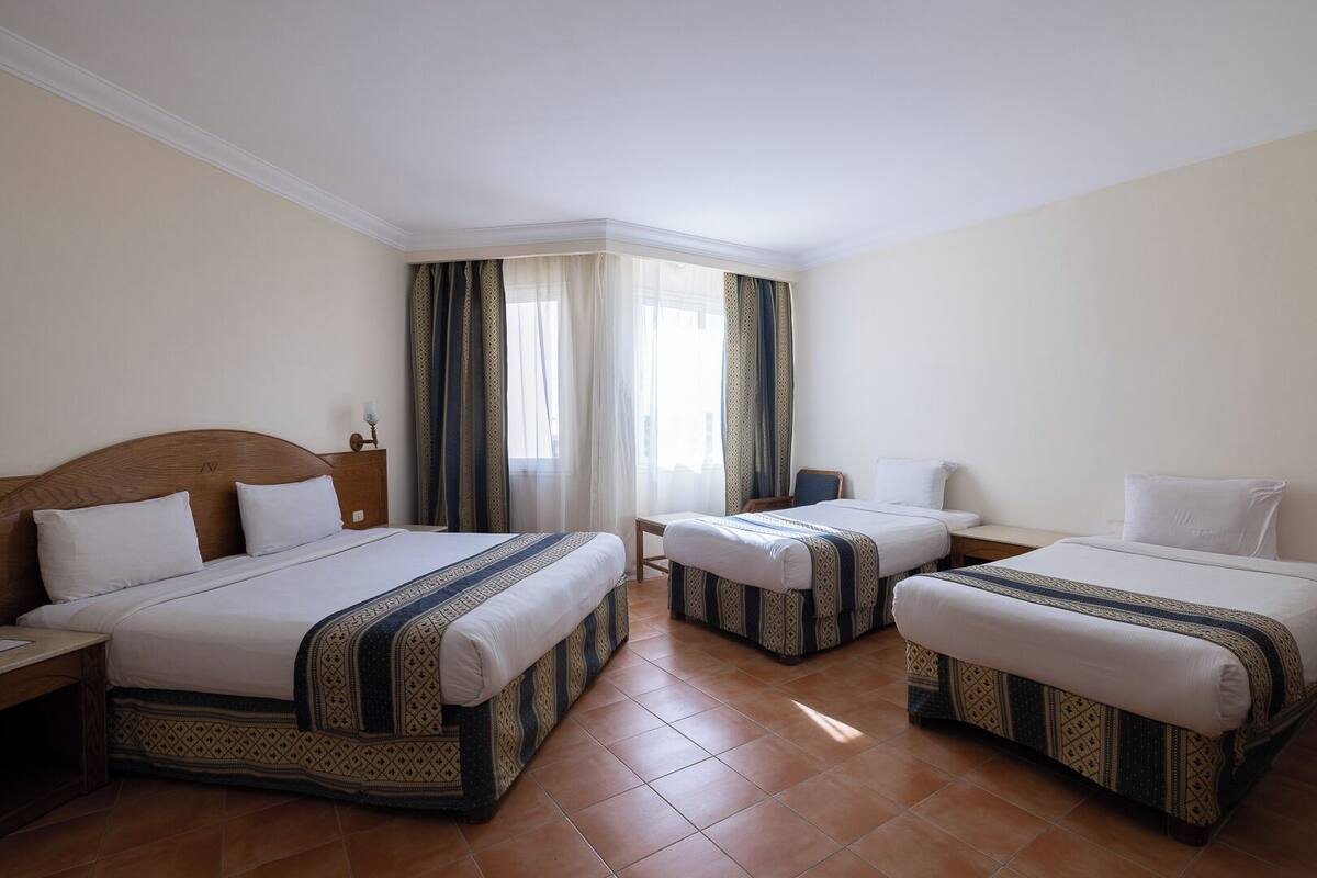 A hotel room with two beds, a wooden headboard, a nightstand, a small table, and a window with curtains.