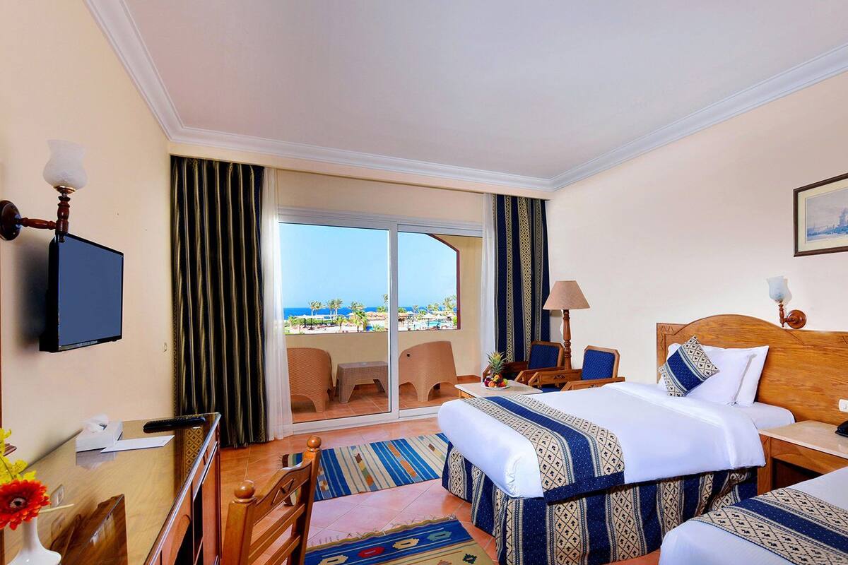 A hotel room with two beds, a television, a desk with a lamp, a chair, and a view of the sea.