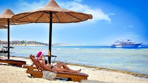 A beach scene with a person relaxing under a straw umbrella, a boat in the background, and a clear view of the sea. - Bliss Nada Beach Resort (Marsa Alam)