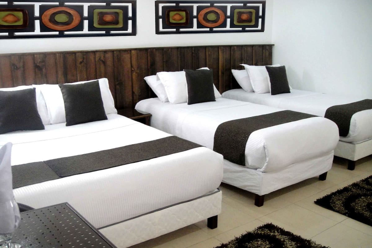 A hotel room with three beds, each with white bedding and black pillows. There is a wooden headboard behind the beds, and the room has a tiled floor.