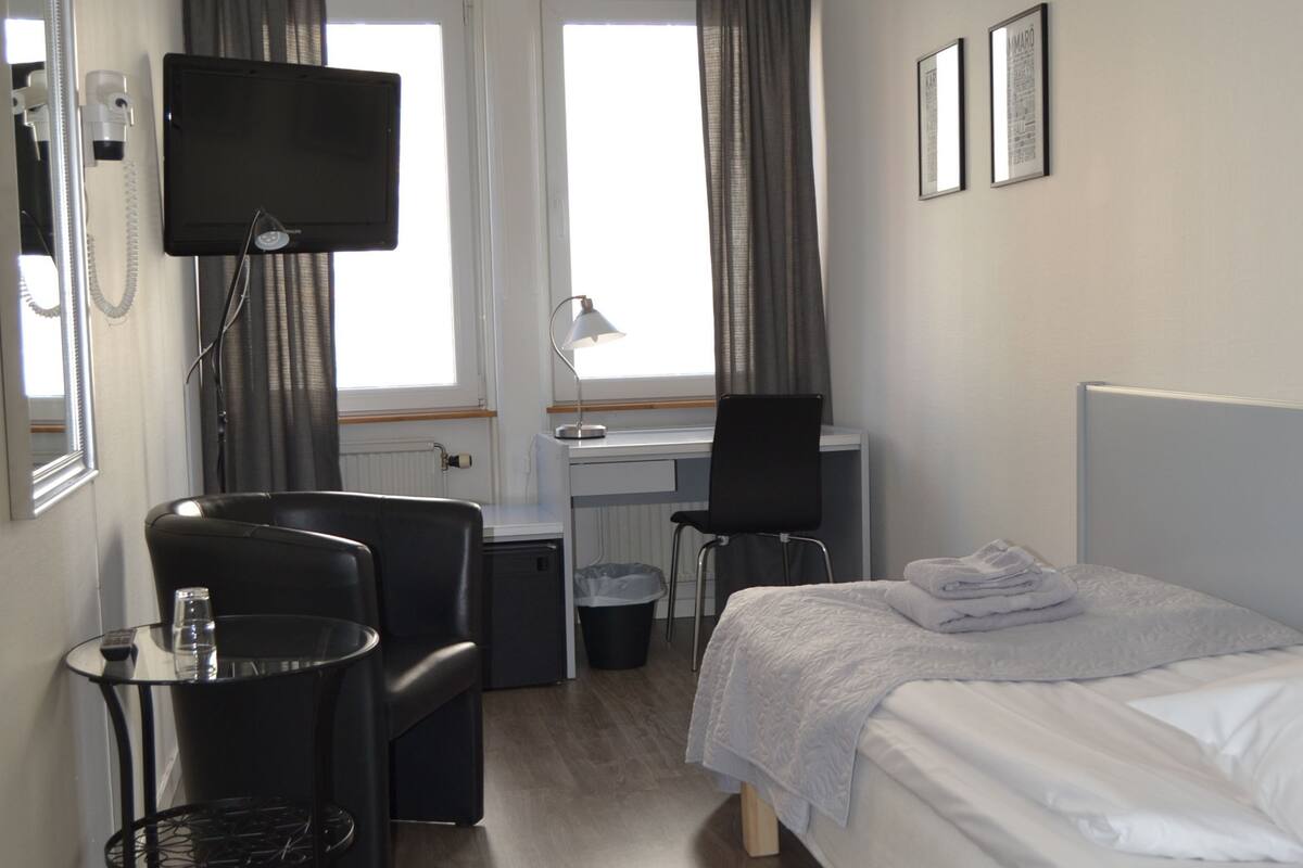 A hotel room with a bed, a desk, a chair, a TV, a small table with a glass, and a trash bin.