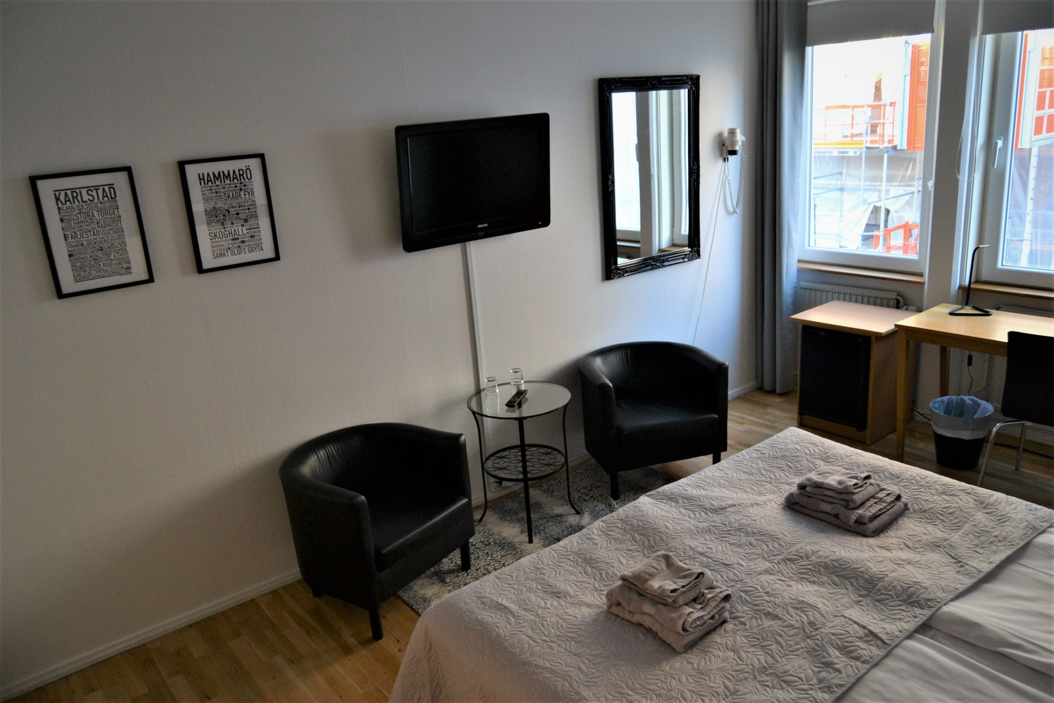 A hotel room with a bed, two black chairs, a small round table, a flat-screen TV, a mirror, and framed pictures on the wall.