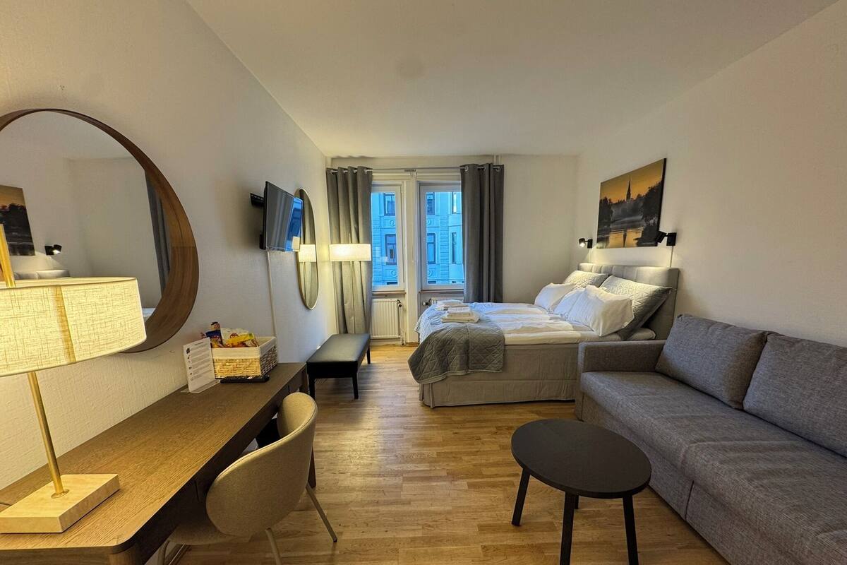 A modern hotel room with a wooden floor, a bed with a grey duvet, a grey sofa, a round table with a lamp, a desk, and a large mirror.