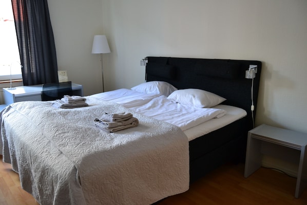 A neatly made bed with white linens and a folded towel on the bed. There is a bedside table with a lamp and a power outlet. The room has a wooden floor and a window with curtains. - Solsta Hotell (Karlstad)