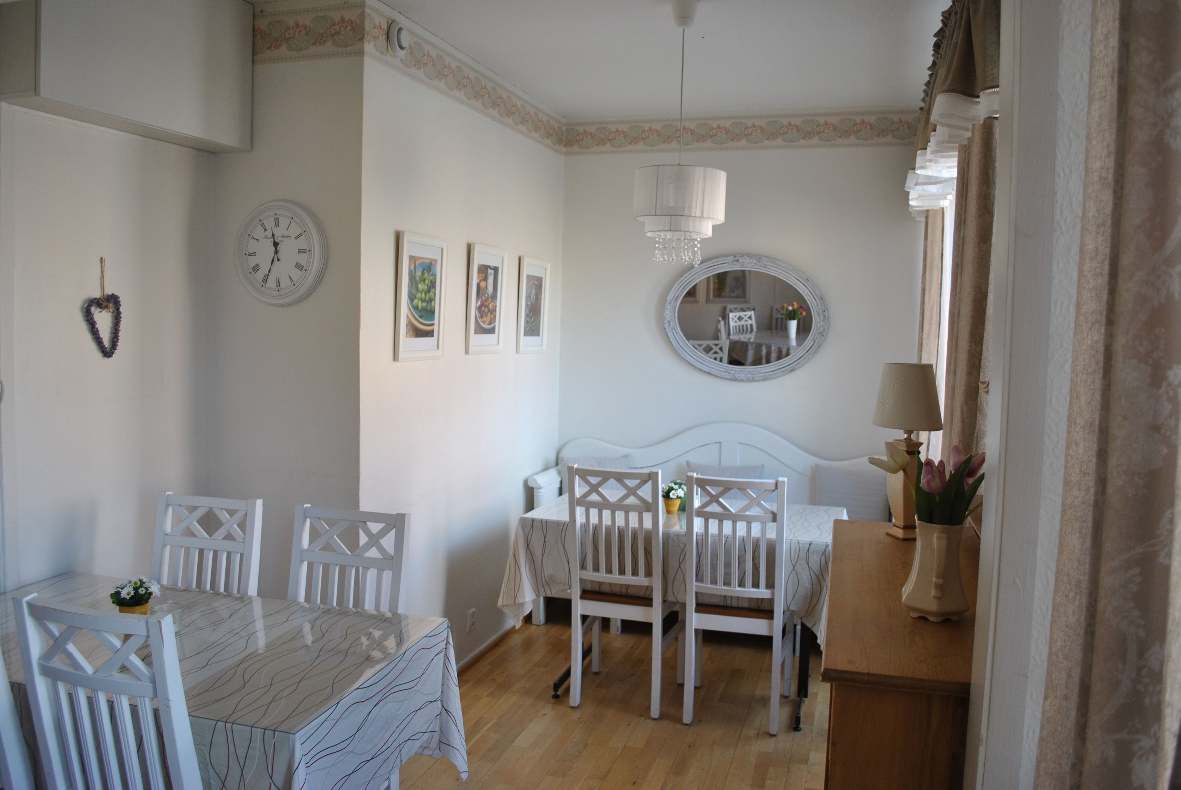 Quarto com mesa de jantar, cadeiras, cama, mesa de cabeceira com abajur, espelho e relógio na parede.