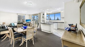A modern living room with a dining area, kitchen, and large windows offering a view of trees and buildings. - The Ritz Resort (Broadbeach)
