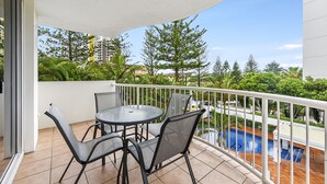 A balcony with a round table and chairs, a pool, and a view of trees and buildings. - The Ritz Resort (Broadbeach)