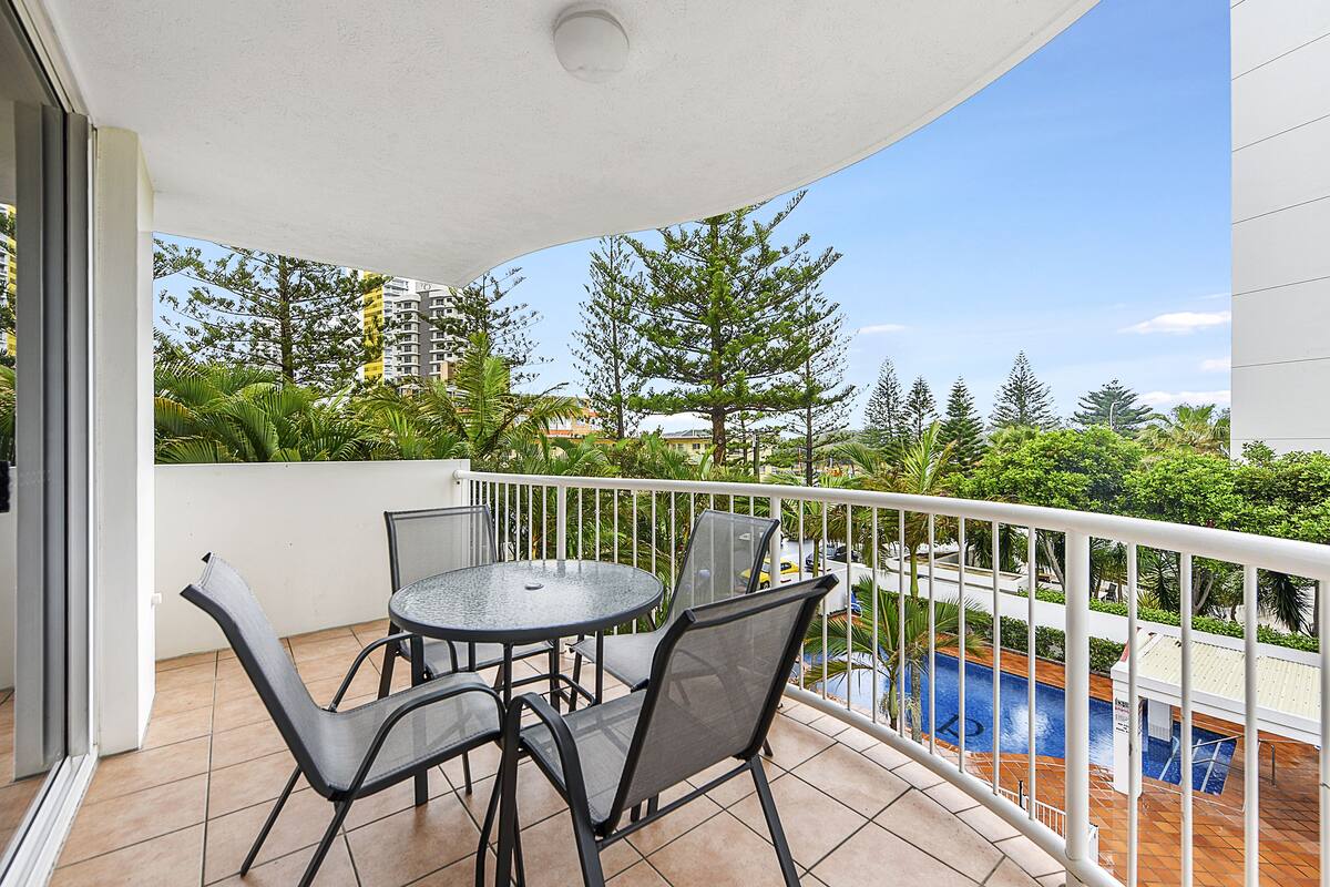 A balcony with a round table and chairs, a pool, and a view of trees and buildings.