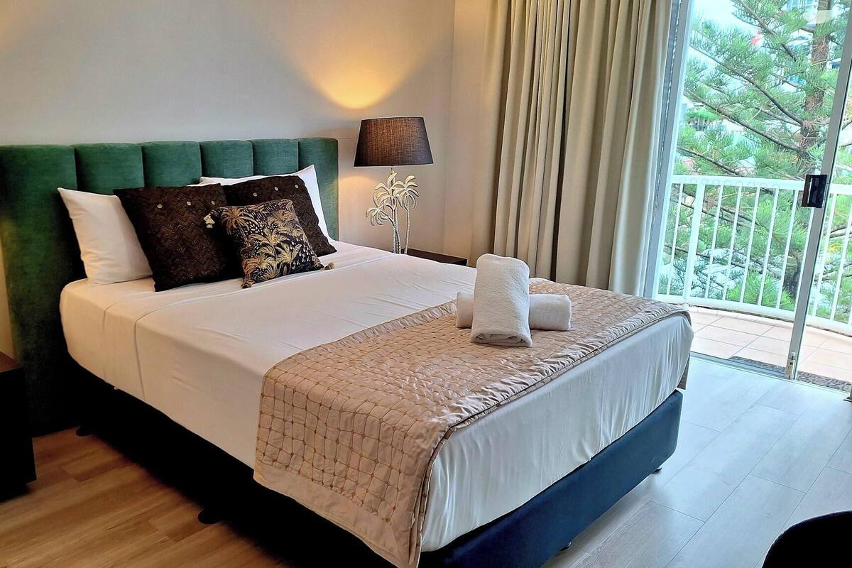 A hotel room with a bed, two bedside tables, a nightstand, a window with curtains, and a view of greenery outside.
