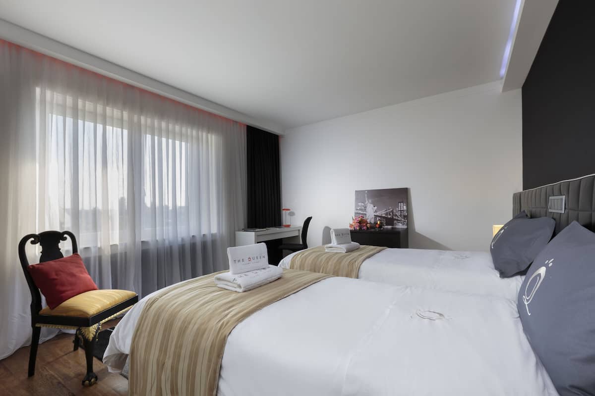 Ein Hotelzimmer mit zwei Betten, einem Schreibtisch, einem Stuhl und einem großen Fenster mit durchsichtigen Vorhängen.