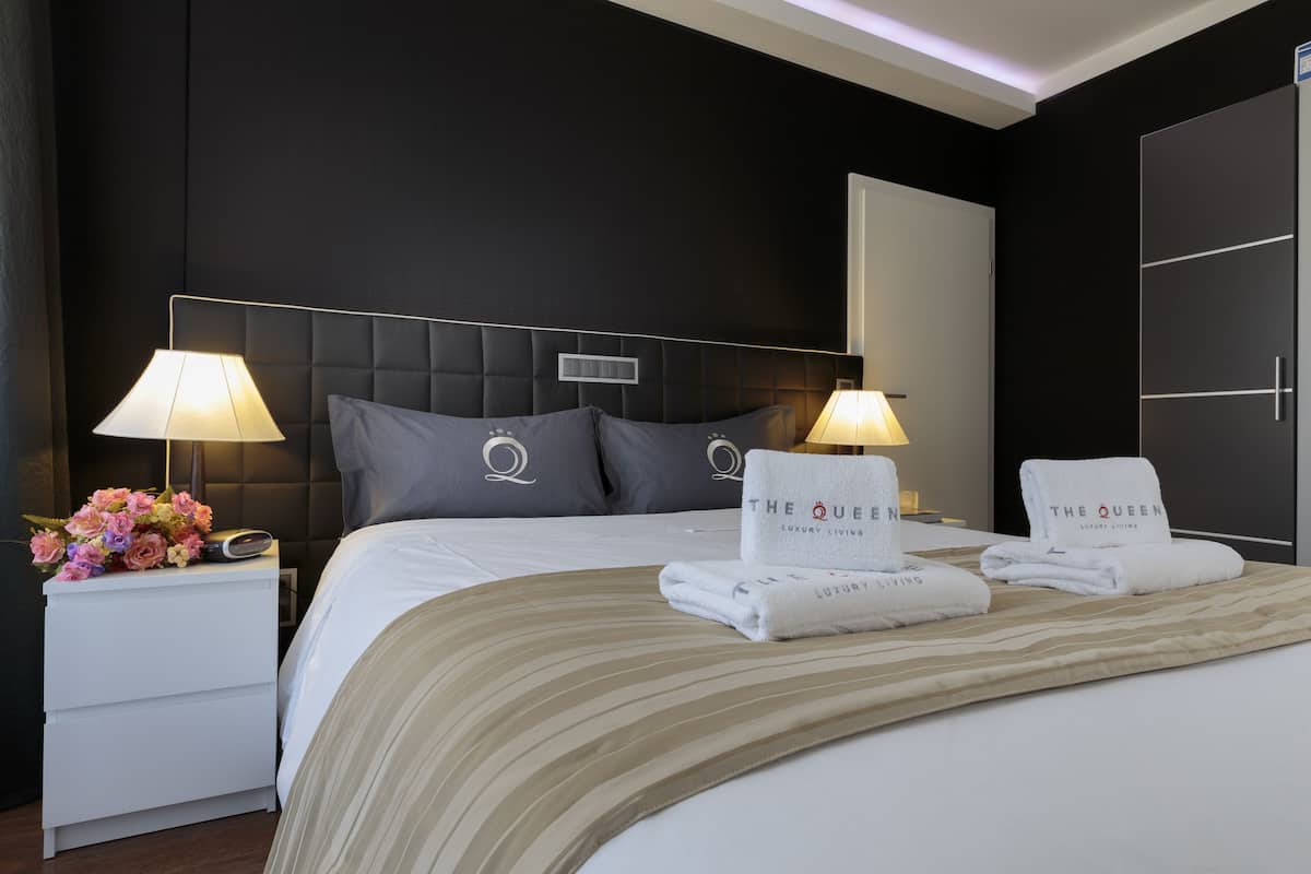 Ein Hotelzimmer mit einem großen Bett, zwei Nachttischlampen, einem Nachttisch mit Blumenarrangement und zwei Handtüchern mit dem Branding "The Queen Luxury Living".