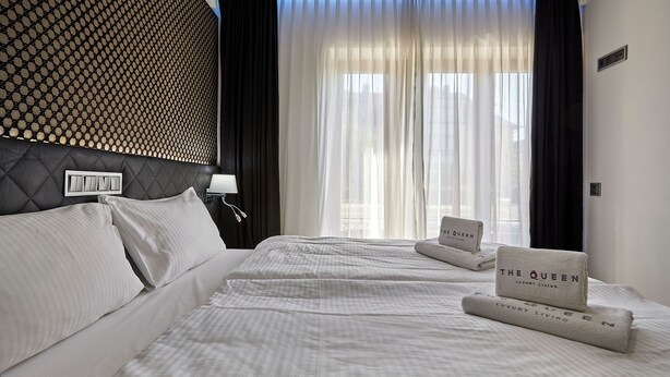 Habitación de hotel con una cama grande, dos almohadas, cabecero con diseño geométrico, mesita de noche con lámpara y ventana con cortinas traslúcidas.
