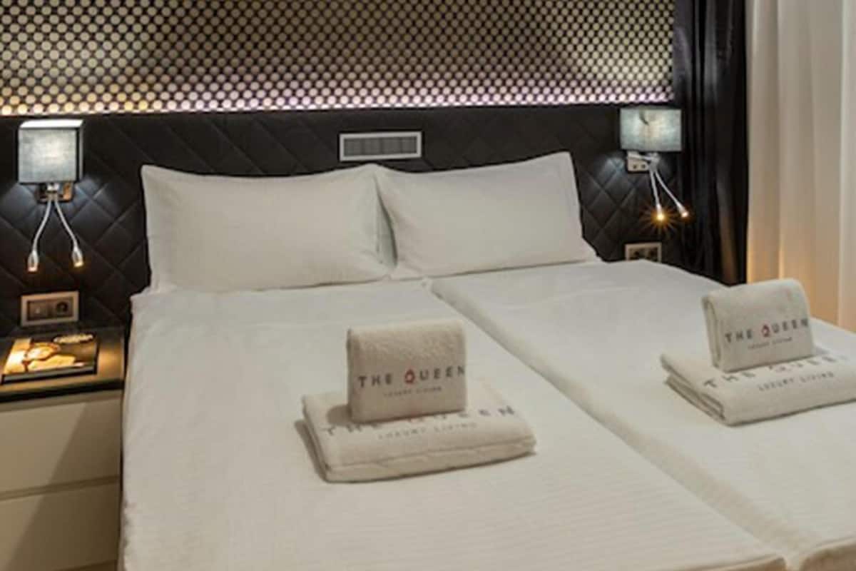 Ein Hotelzimmer mit einem großen Bett, zwei Kissen, einem Nachttisch mit Lampe und zwei Handtüchern mit dem Schriftzug 'The Queen'.