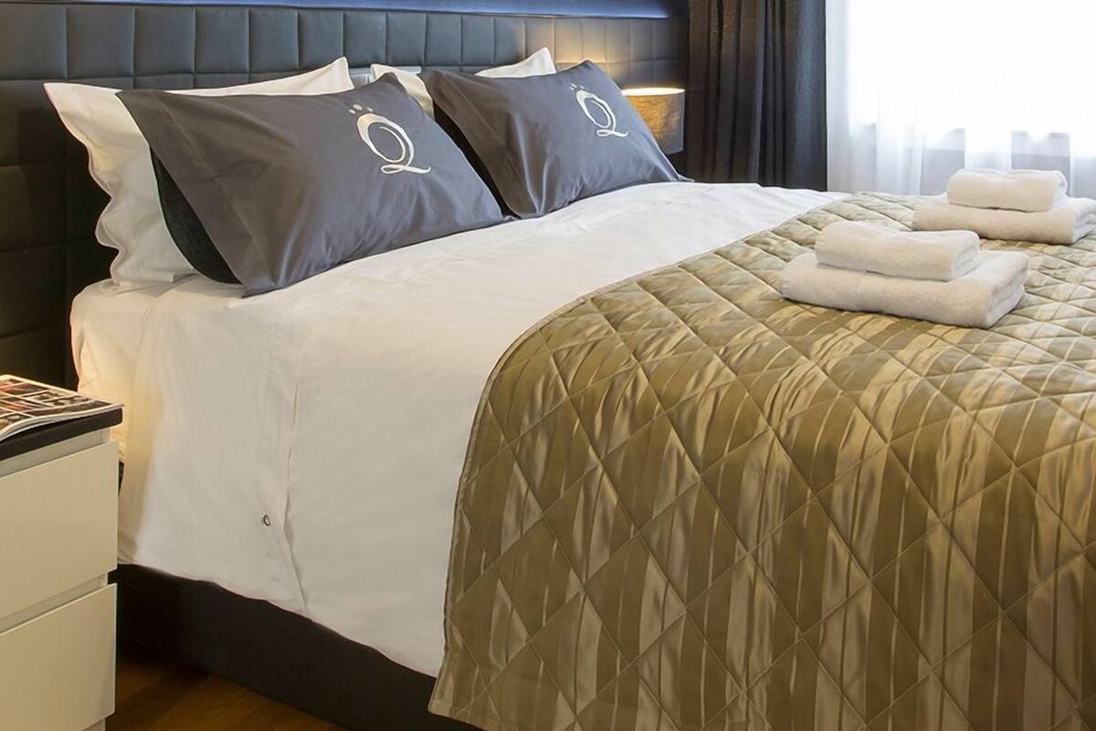 Uma cama bem arrumada com cobertura acolchoada dourada, duas almofadas com um logotipo e uma mesa de cabeceira com uma revista.