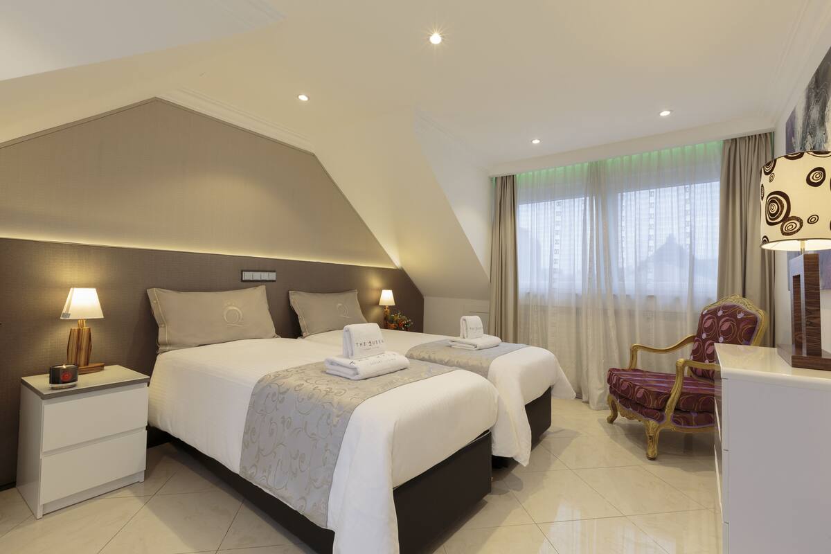 Quarto de hotel moderno com duas camas, uma mesa de cabeceira com abajur, uma cadeira e uma escrivaninha com luminária.