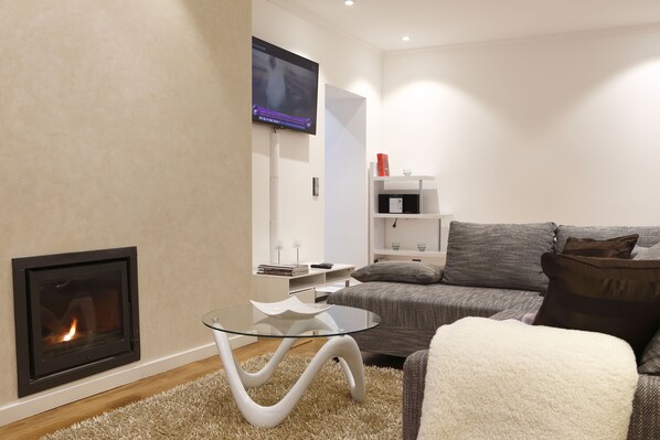 A modern living room with a fireplace, a glass coffee table, a grey sofa, and a flat-screen TV mounted on the wall.