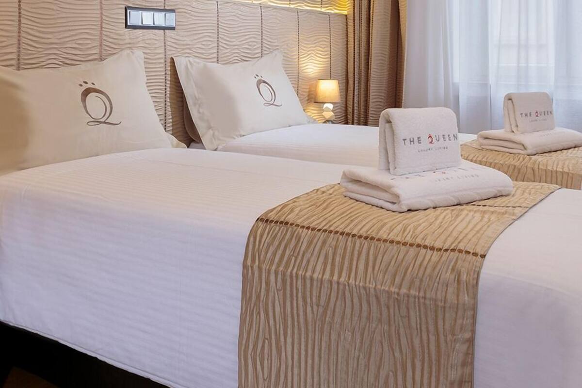 Quarto de hotel com duas camas, cada uma com cabeceira bege e almofadas a combinar. Há uma toalha dobrada com a marca 'The Queen' na cama.