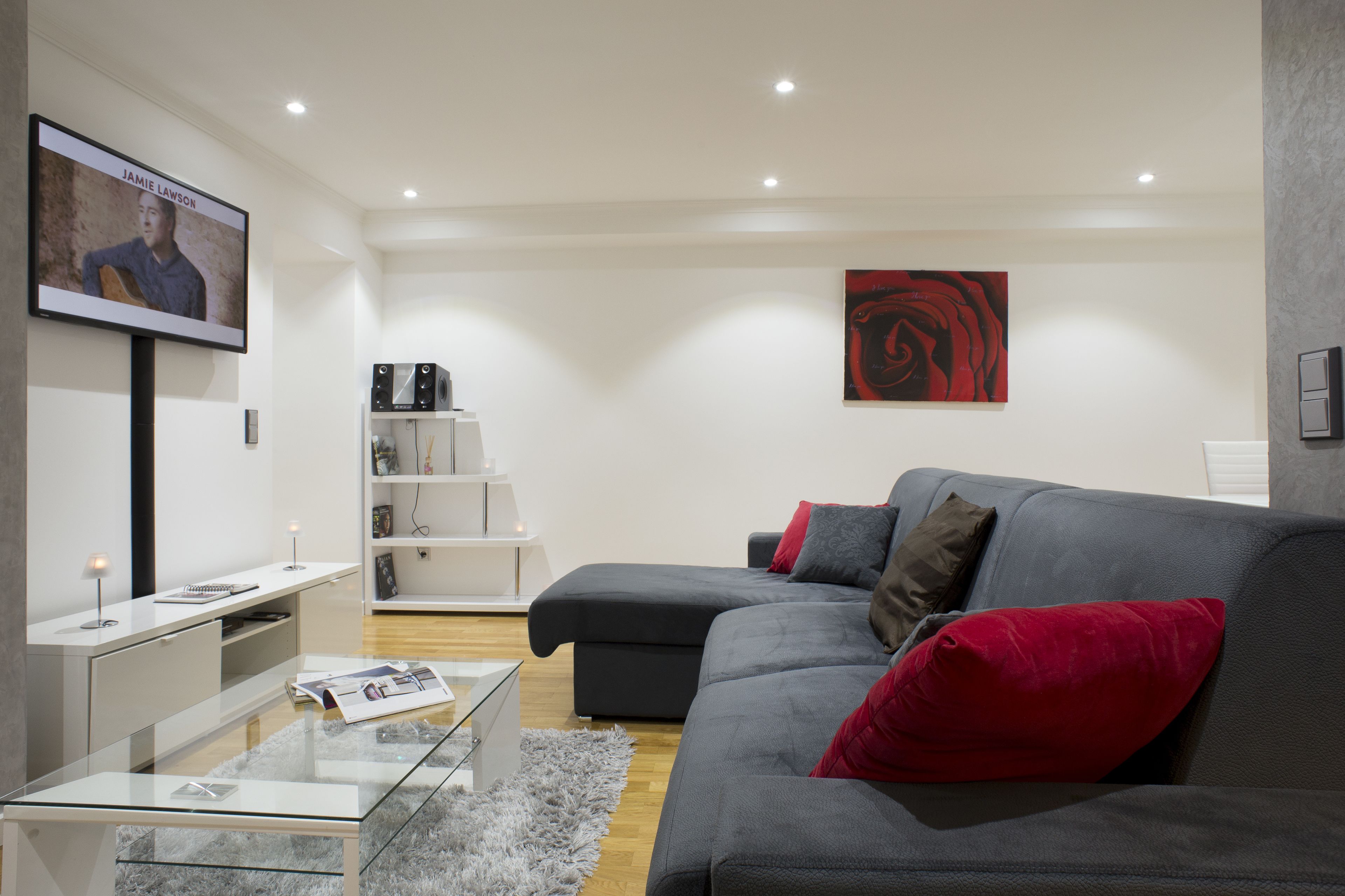 A modern living room with a grey sofa, red and brown cushions, a glass coffee table, a white entertainment unit, and a red abstract painting on the wall.