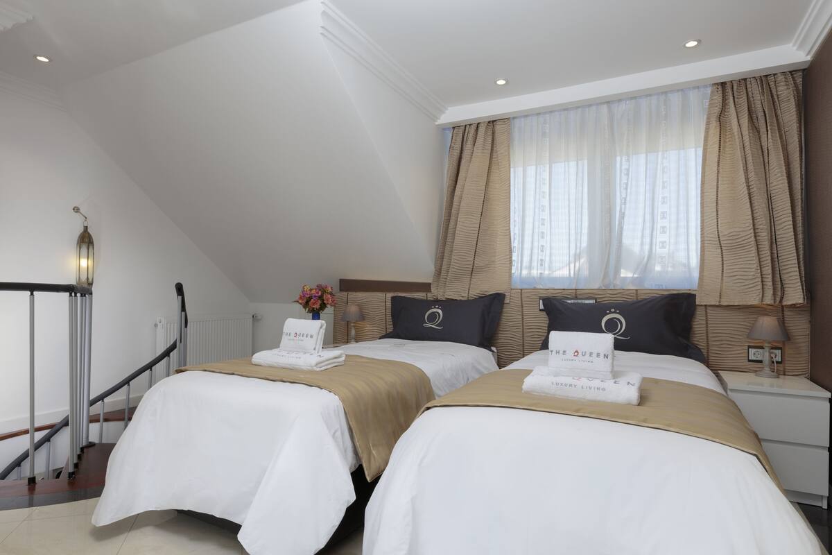 Quarto de hotel com duas camas, uma mesa de cabeceira com abajur, uma janela com cortinas finas e um aviso na cama que diz "The Queen Luxury Living".