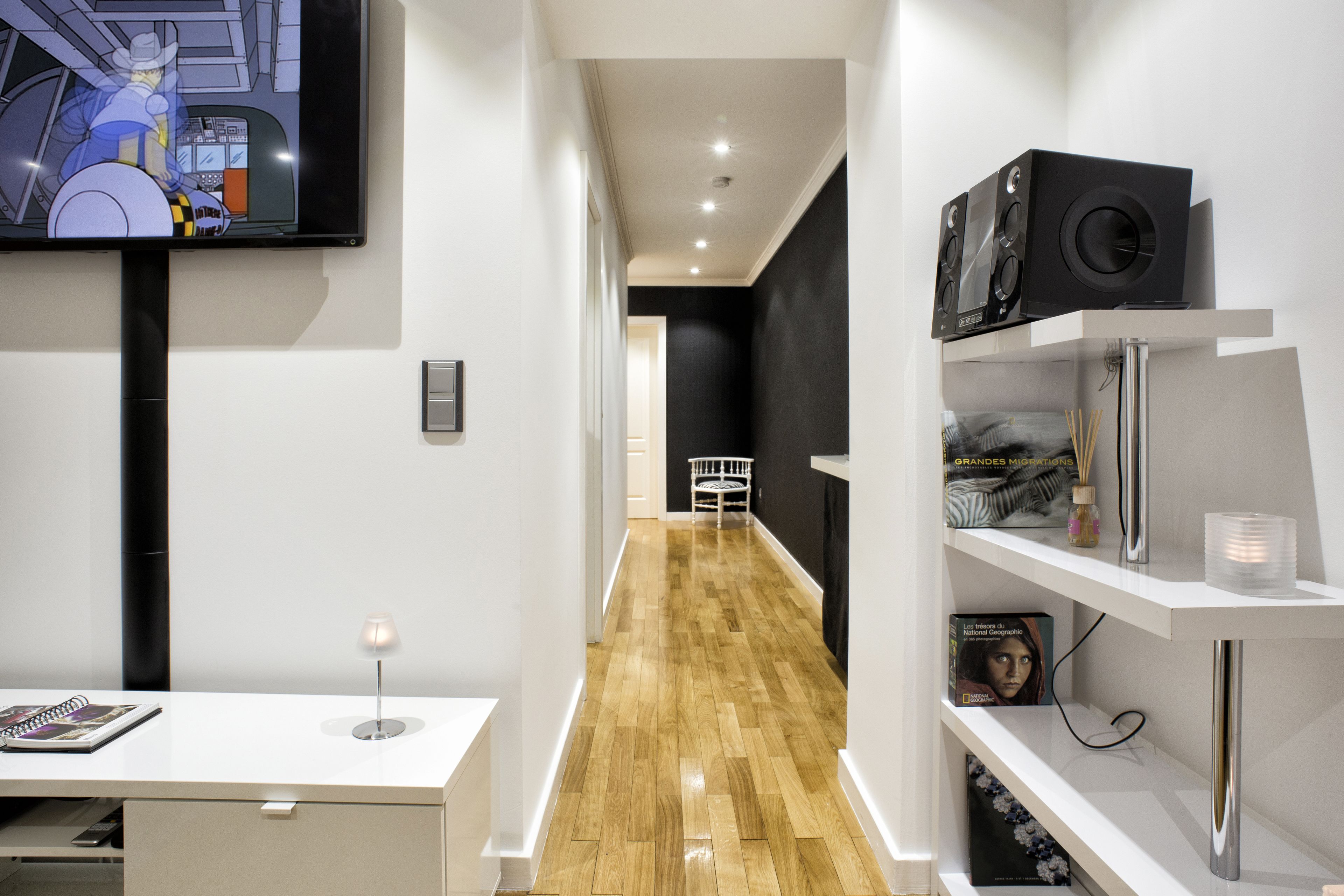 A modern hotel corridor with a wooden floor, a TV mounted on the wall, a desk with a lamp, and a shelf with books and a speaker.