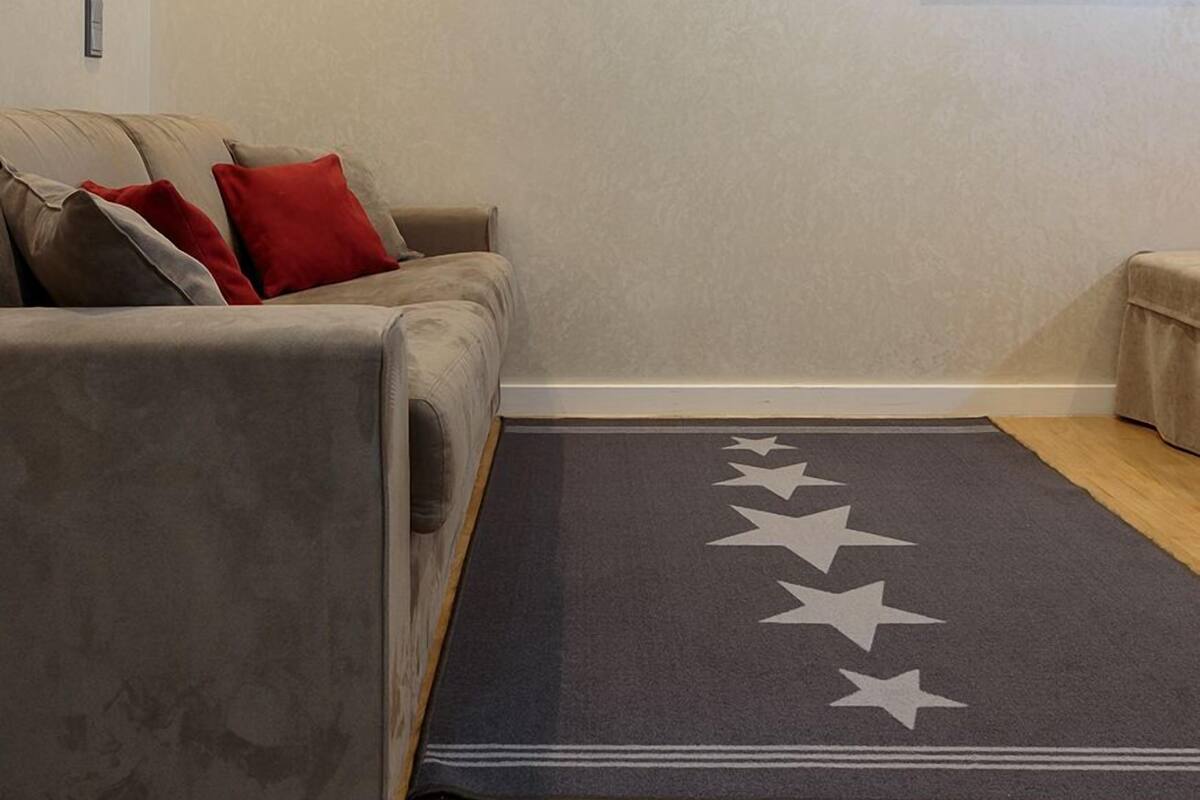 Sala de estar com um sofá cinza, almofadas vermelhas e cinzas, e um tapete escuro com estrelas brancas.