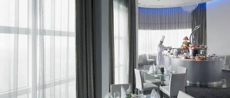 A modern hotel dining area with a chef preparing food, large windows with sheer curtains, and a buffet setup.