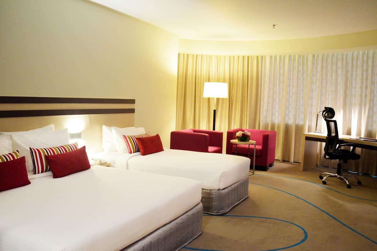A hotel room with two beds, a desk, a chair, and a lamp.