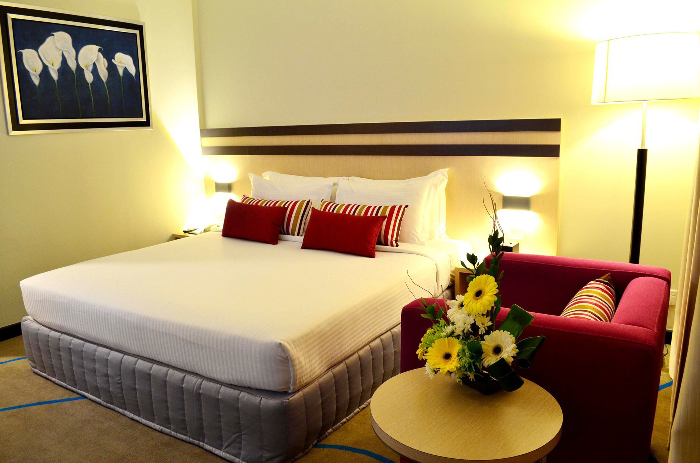 A hotel room with a large bed, a red armchair, a small round table with a vase of flowers, and a painting on the wall.