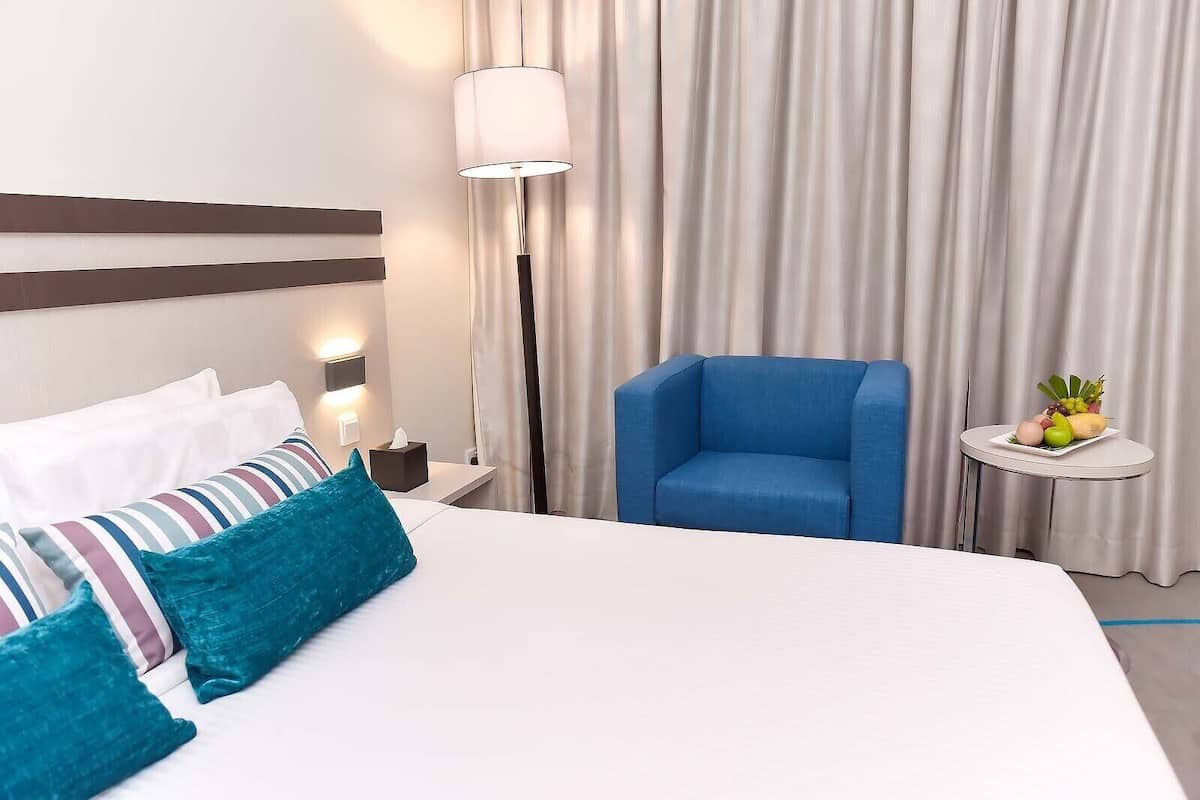 A hotel room with a bed, a blue armchair, a bedside table with a fruit plate, and a lamp.