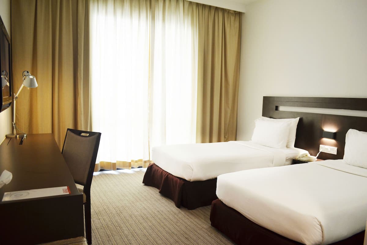 A hotel room with two beds, a desk, a lamp, and a large window with curtains.